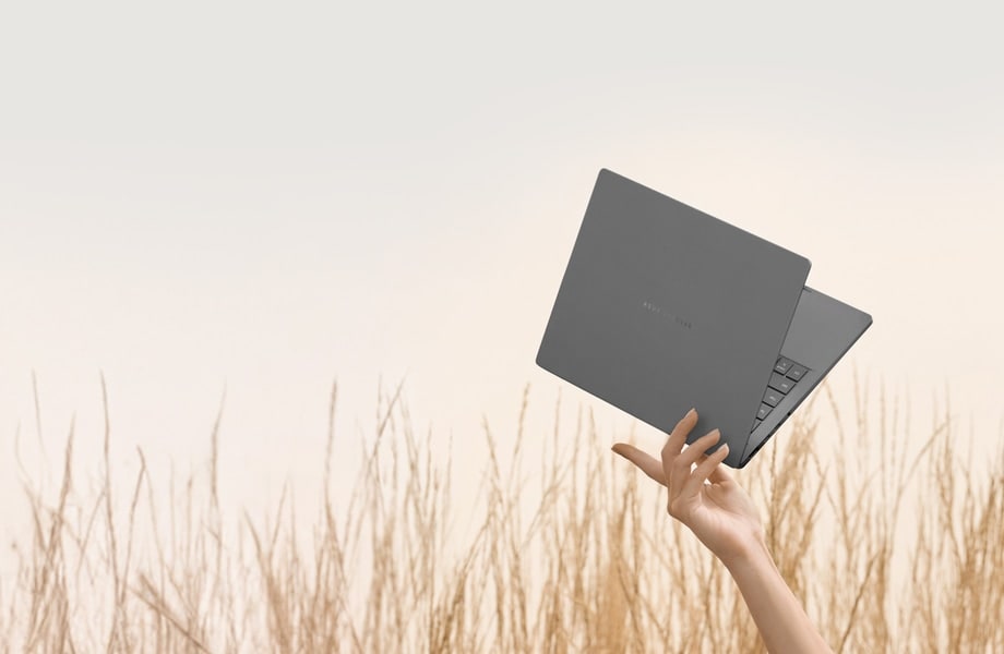 A gray Zenbook A14 is easily lifted with just a few fingers. The computer is slightly open at about a 40-degree angle, facing toward the upper right.