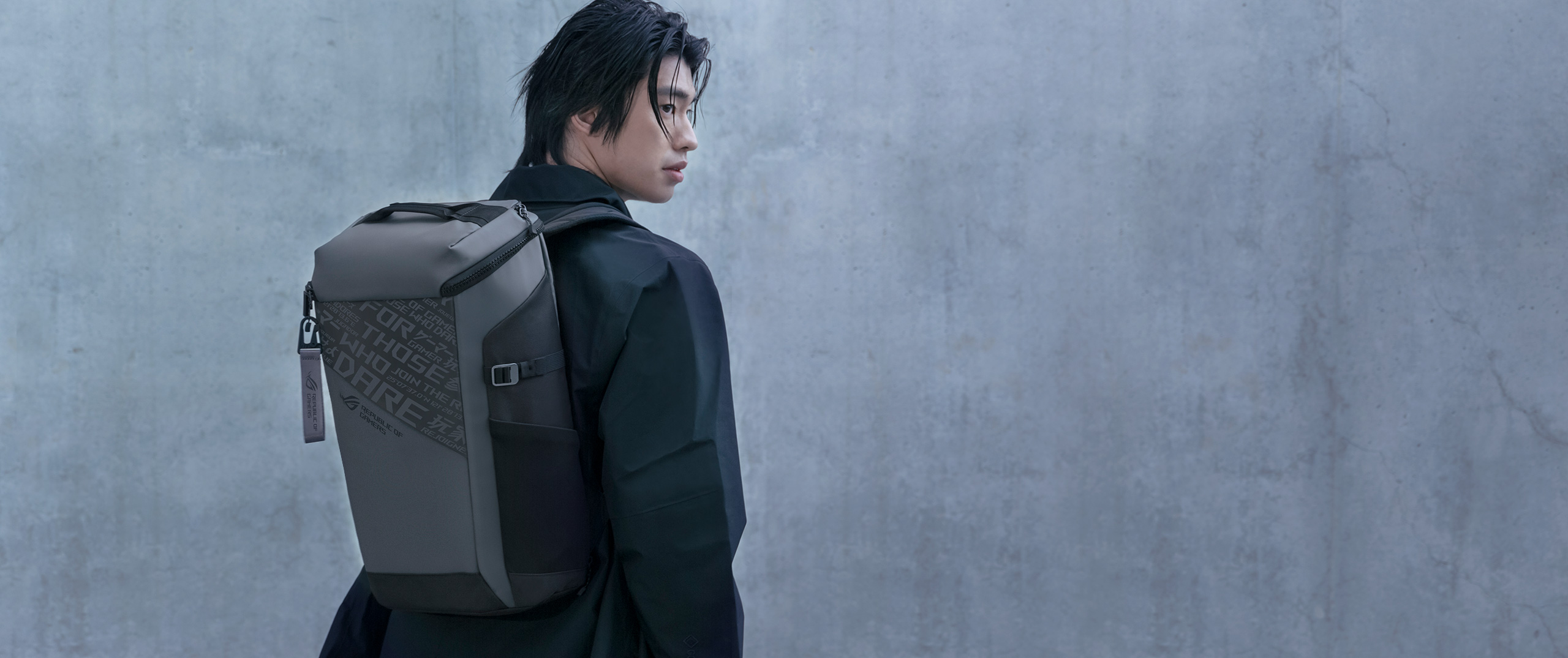 A person with a dark jacket standing near a concrete wall, looking over his shoulder and wearing an ROG Ranger BP2701 Gaming Backpack.