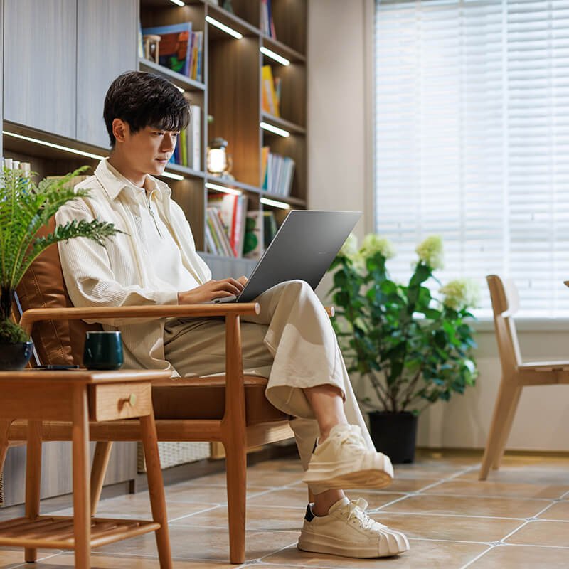 A young man is sitting in a chair using ASUS Vivobook S16 with several plants in the background.
