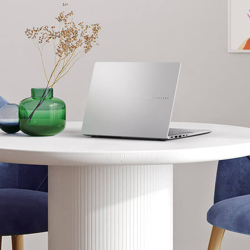 A silver ASUS Vivobook S14 sits on a modern-looking white table with two blue chairs on either side of it.