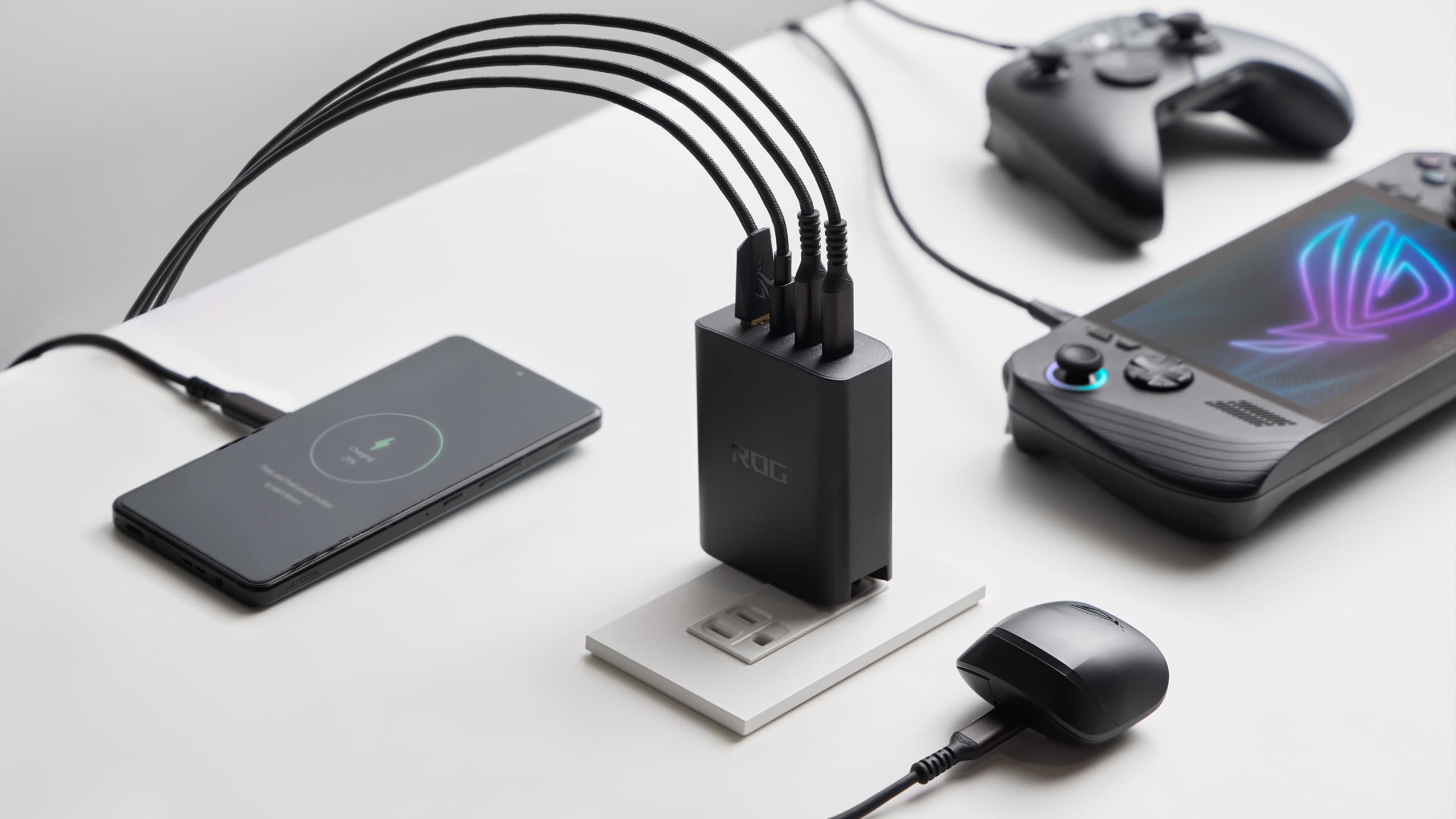 An ROG galium-nitride charged plugged into a wall socket, with all four ports occupied and charging a phone, ROG Ally, gamepad, and wireless earbuds.